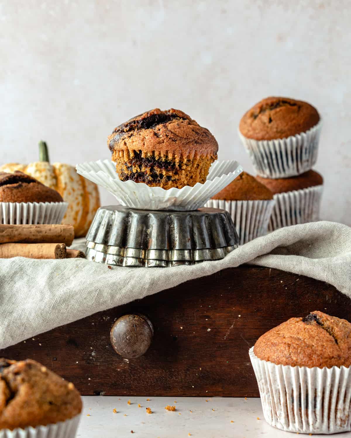 Muffins on pastry case surrounded with other muffins and a pumpkin with a grey cloth