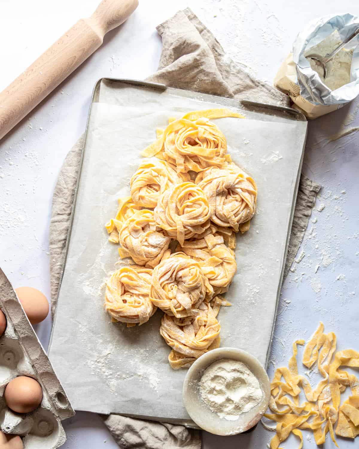 Nest of homemade pasta on a tray with eggs and a rolling pin with a bowl of flour