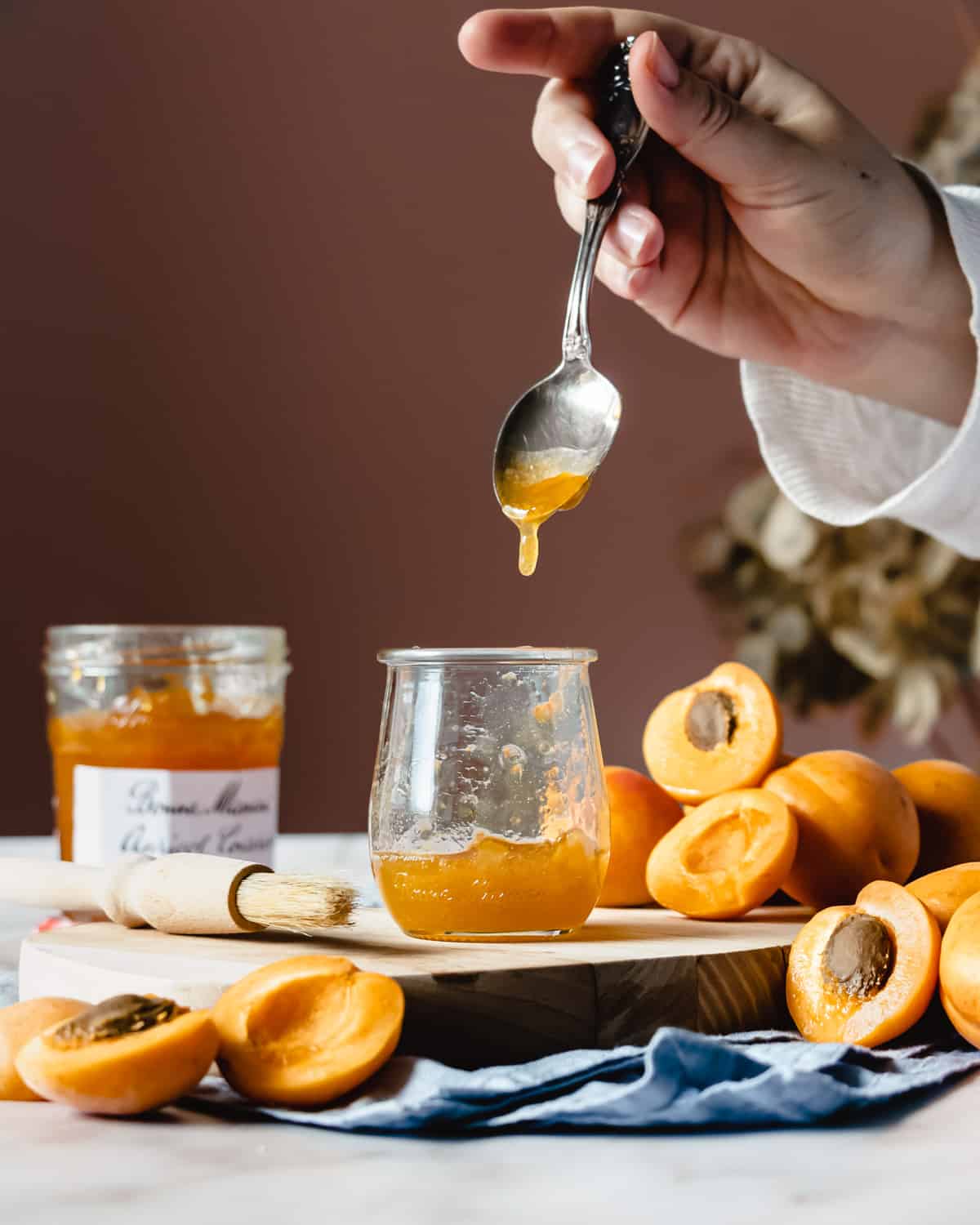 Poring apricot jam into a pot glass on a wooden chopping board with apricot around. 