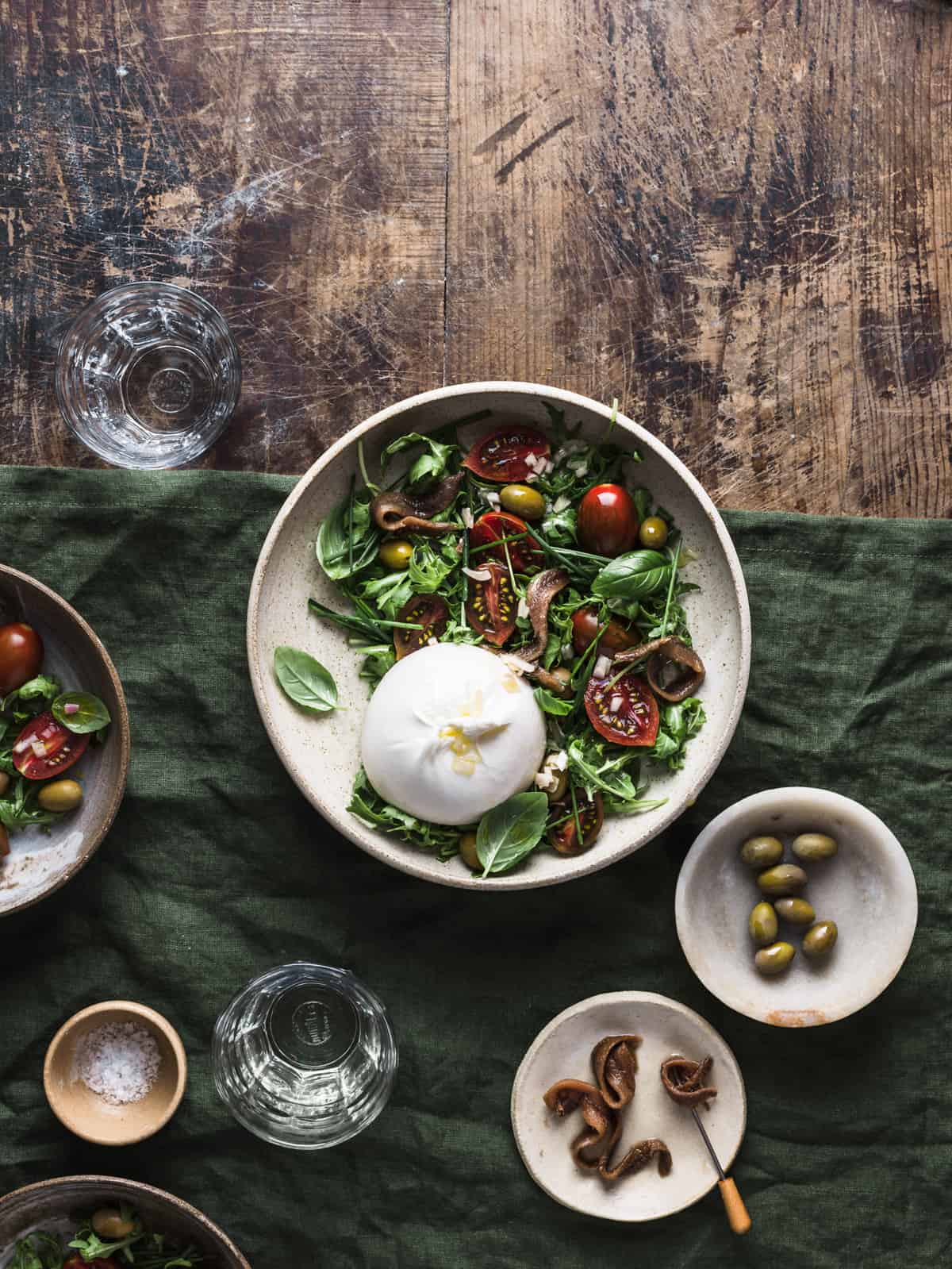 Overhead shot showcasing a salad bowl full of burrata, arugula, tomatoes, olives, and anchovies on a green linen tablecloth with other bowls and glasses around.