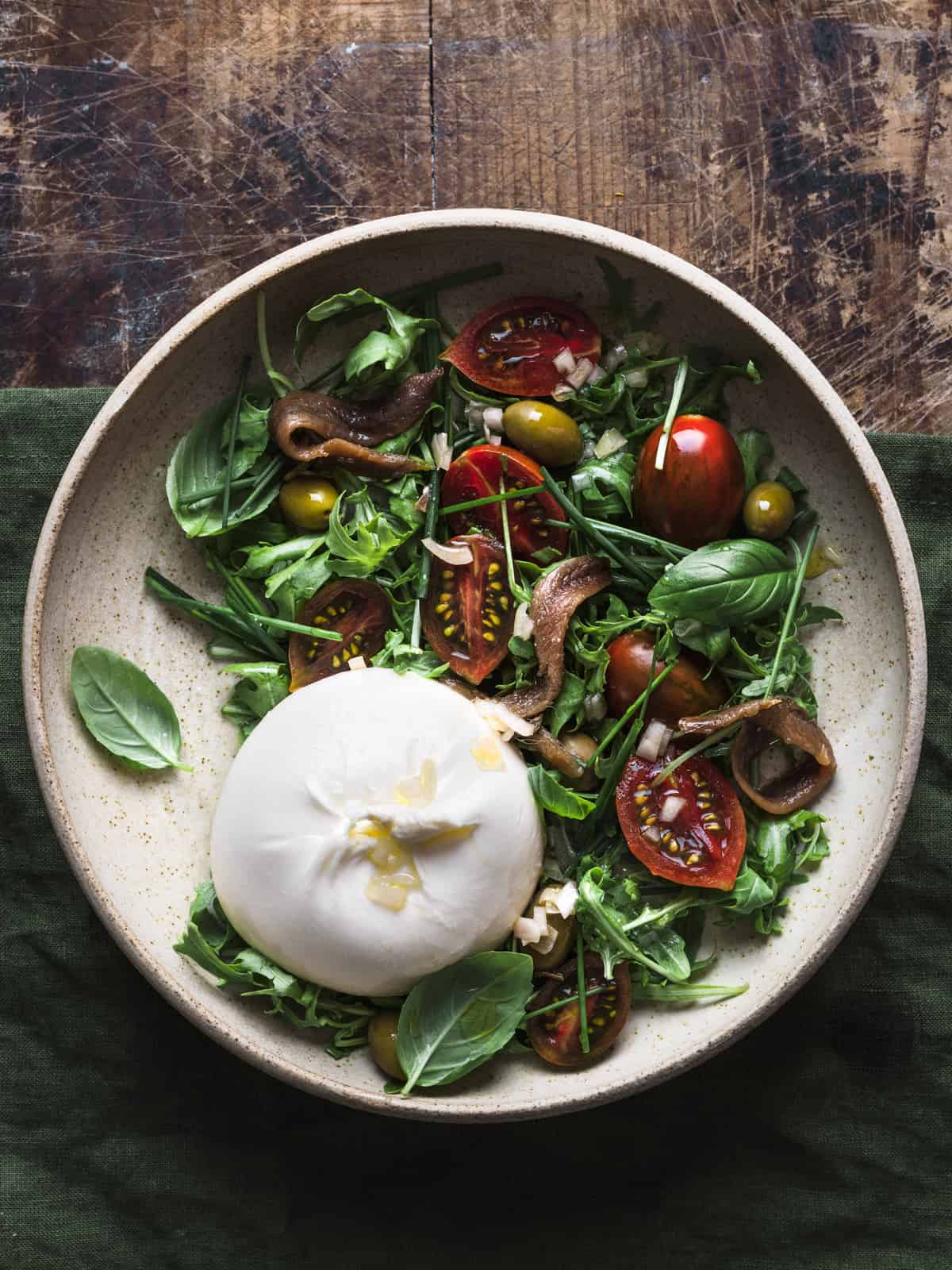 Overhead shot showcasing a salad bowl full of burrata, arugula, tomatoes, olives, and anchovies on a green linen tablecloth.