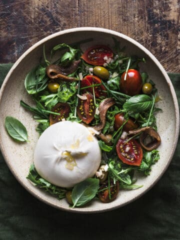 Bowl full of salad with burrata, tomatoes, arugula, anchovies and olives on a green linen tablecloth.