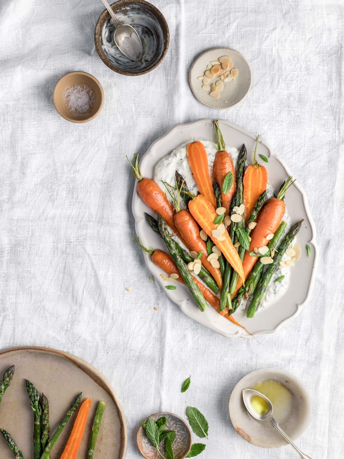 Overhead shot of serving plate with carrots and asparagus on a white table surrounded by littles bowls full of oil, mint leaves and yogurt dip.