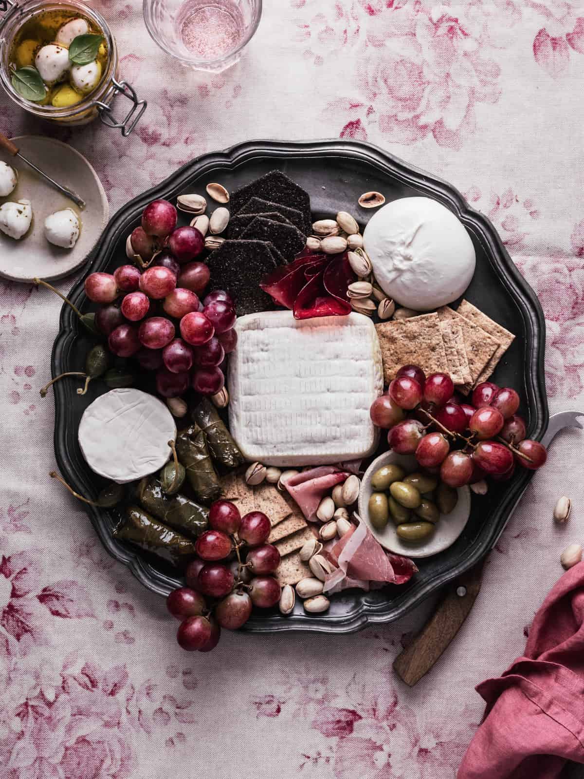 Mediterranean charcuterie board with a cheese knife next to it and on a tablecloth with a jar full of marinated mini mozzarella balls