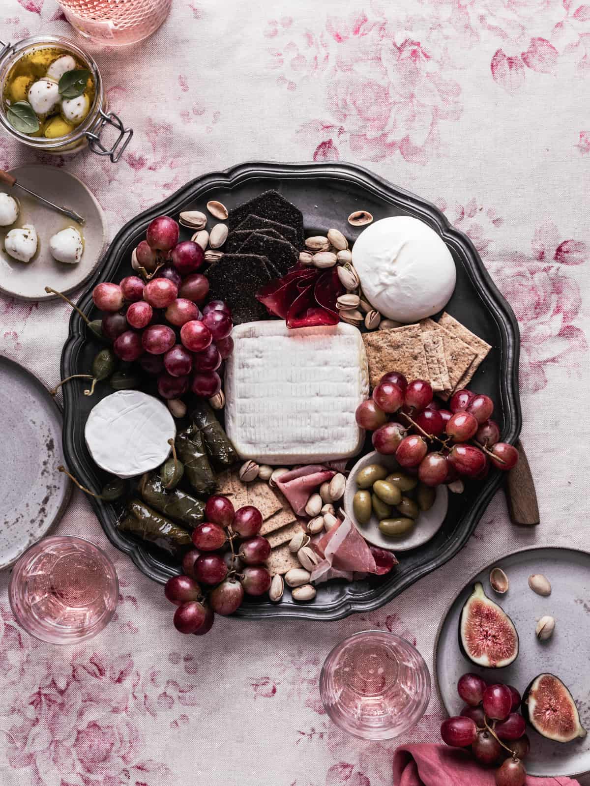 Mediterranean charcuterie board to it and on a tablecloth with a jar full of marinated mini mozzarella balls and few plates on the side with grapes and figs.