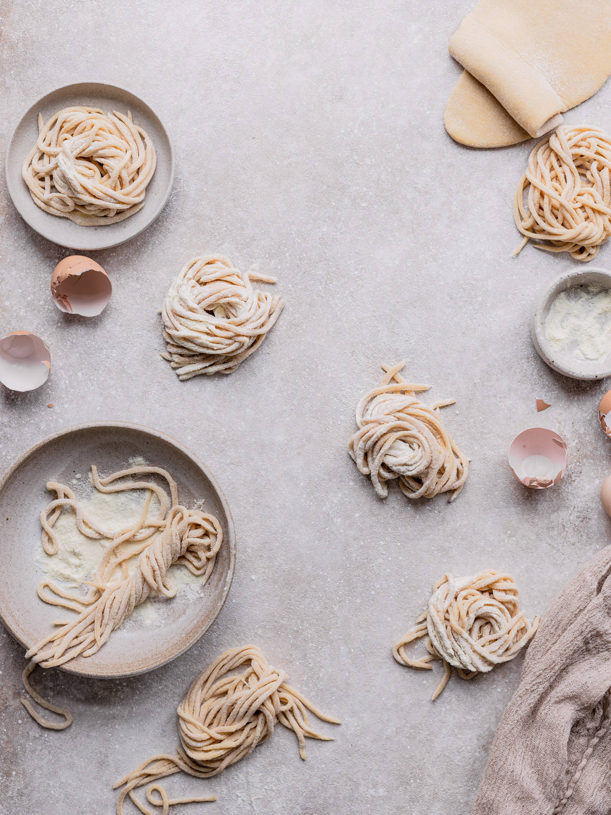 Some nest of tonnarelli pasta on a backdrop and in a bowl with some semolina flour with some eggs shells around