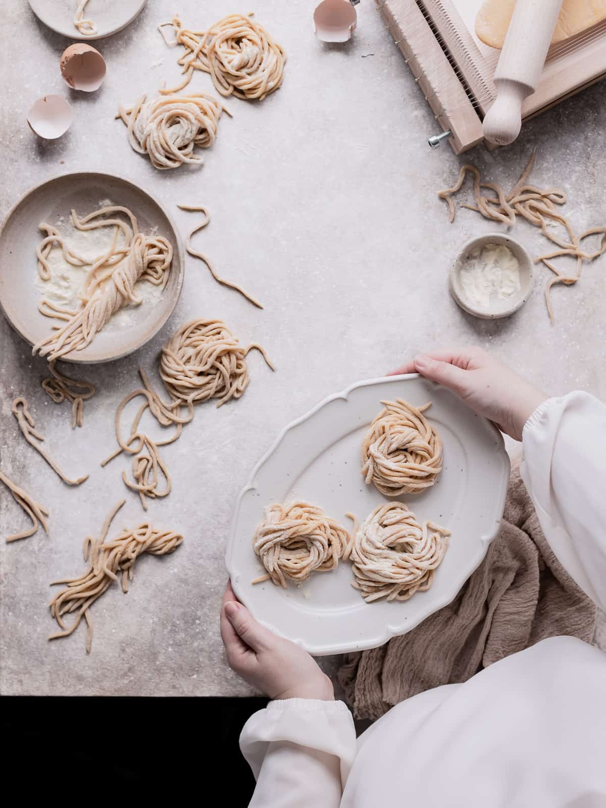 Hands holding a serving dish with some tonnarelli nests on it, while the backdrops has more pasta nest and chitarra toll pasta around the hands.