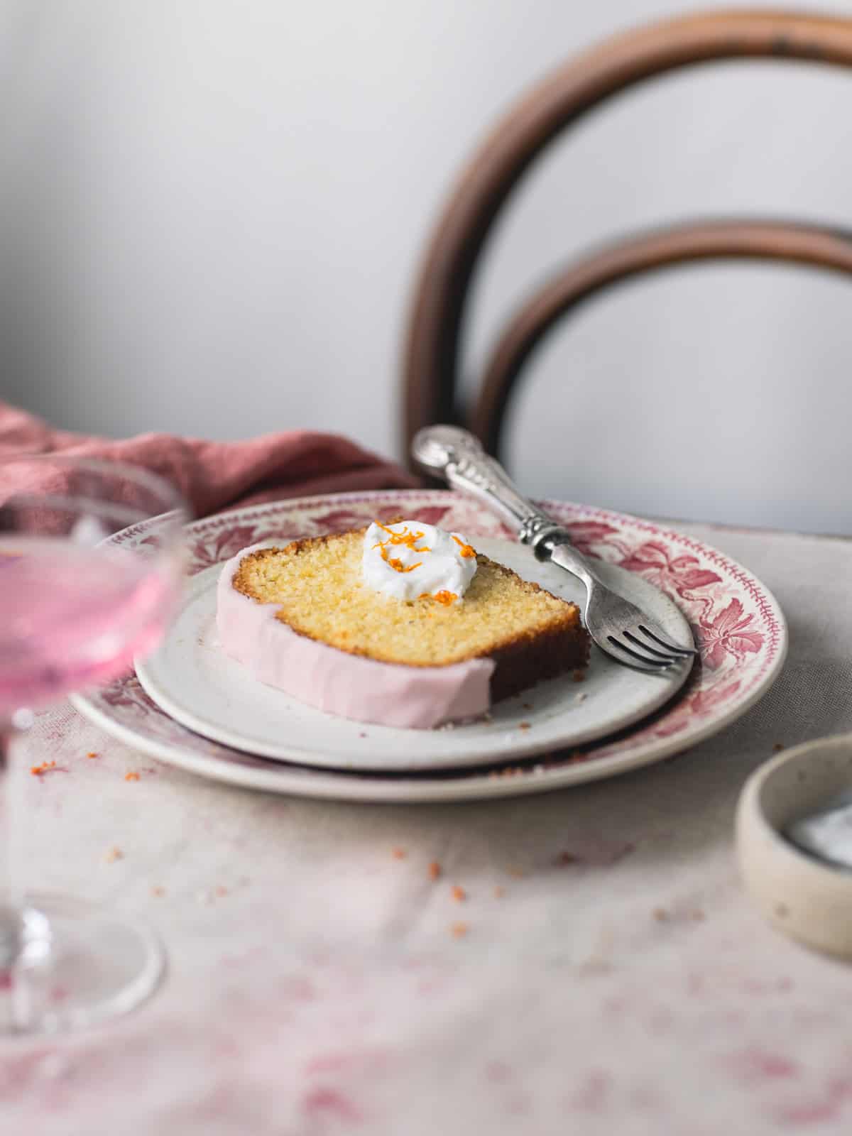 Side photo of a slice of gluten free cake, with on top some whipped cream and few oranges zest, on white plate on a flower background