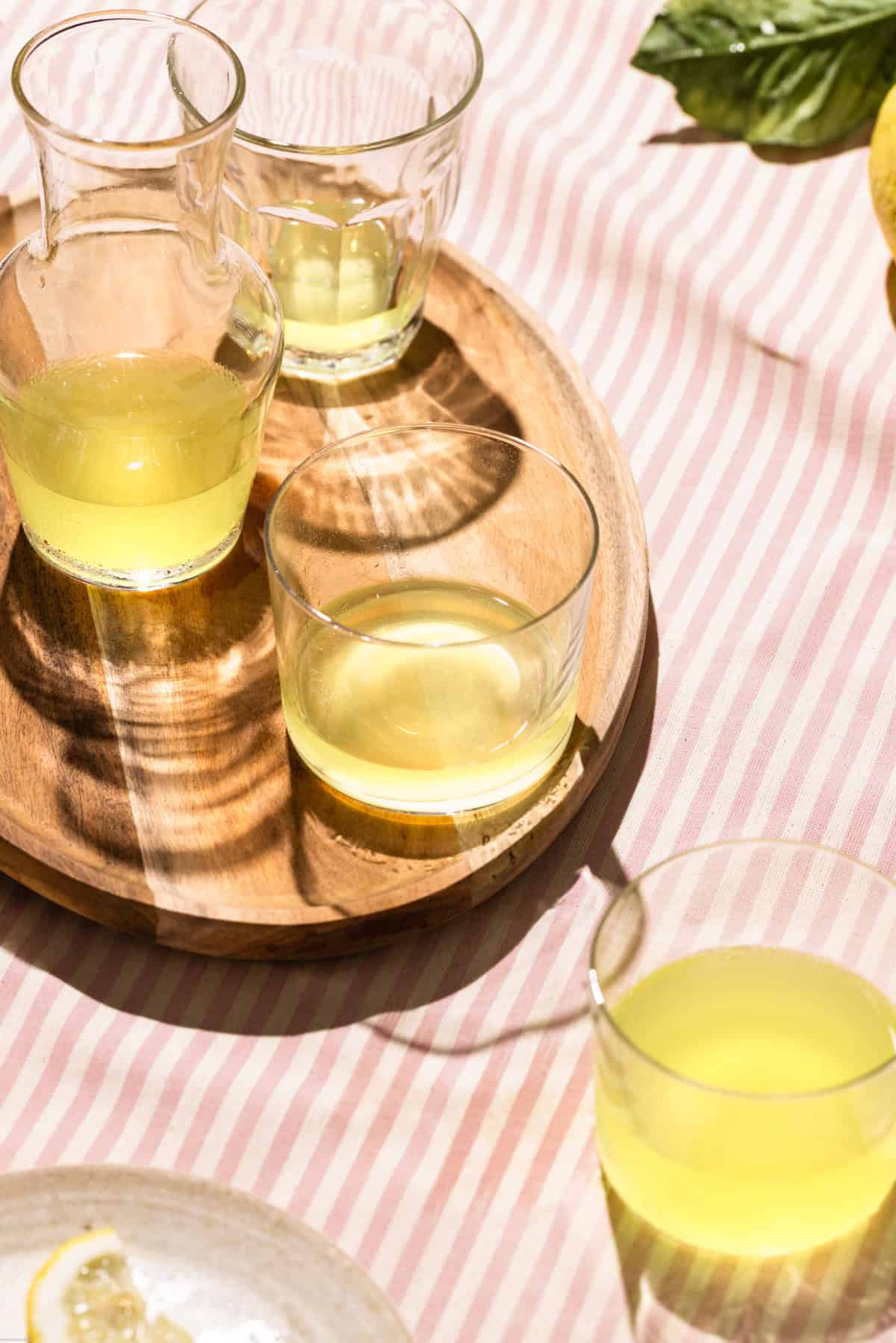 Glasses full of limoncello on a pink stripe tablecloth next to a plate with a slice of lemon.
