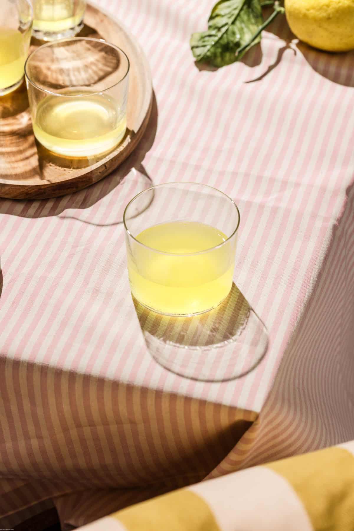 Glasses full of limoncello on a pink stripe tablecloth near a one stripe pillow.