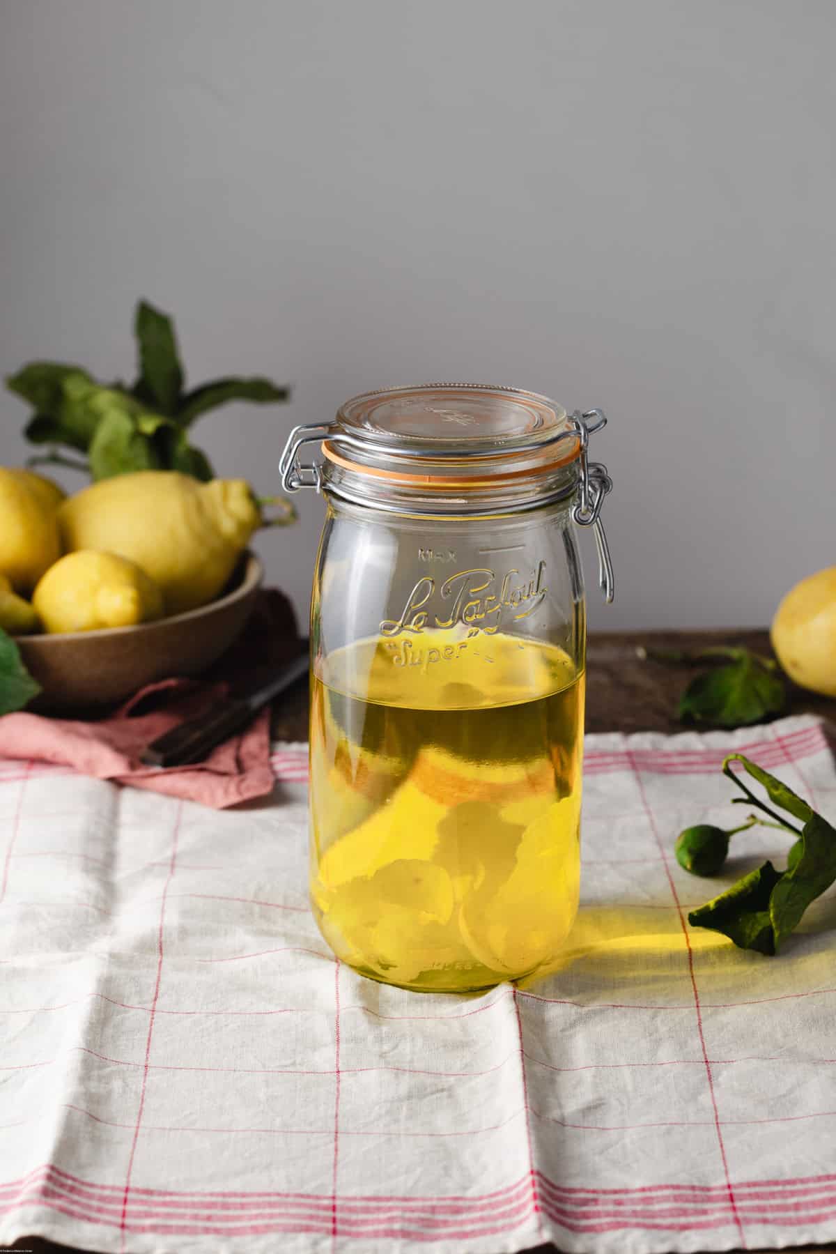 Tall jar full of lemon peel infused placed on a white kitchen towel with lemons around.