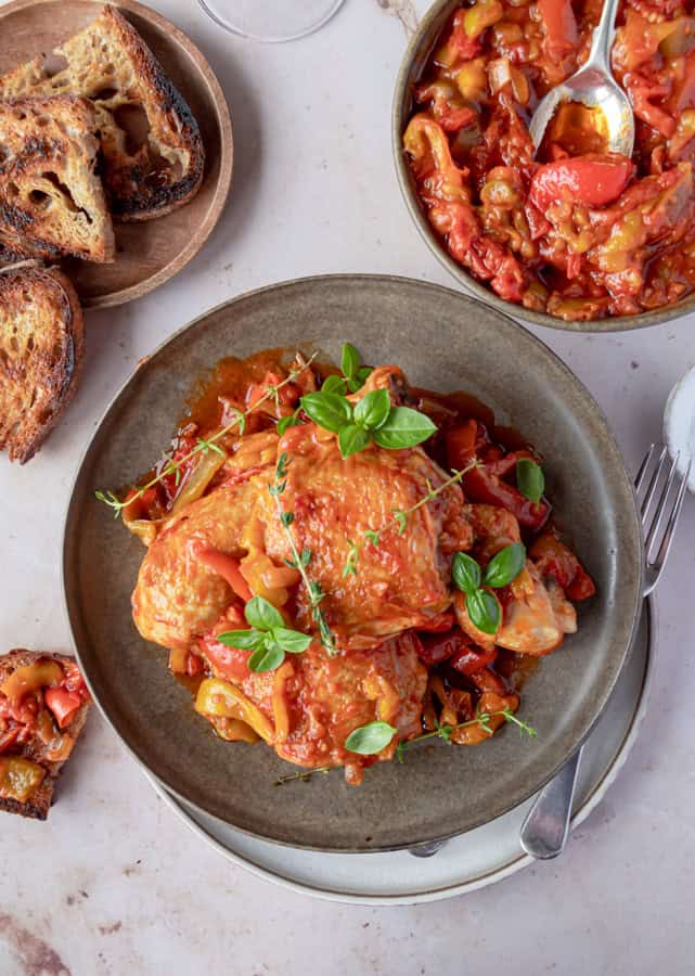 A plate of chicken alla romana with peppers and tomato sauce next to a slice of bread.