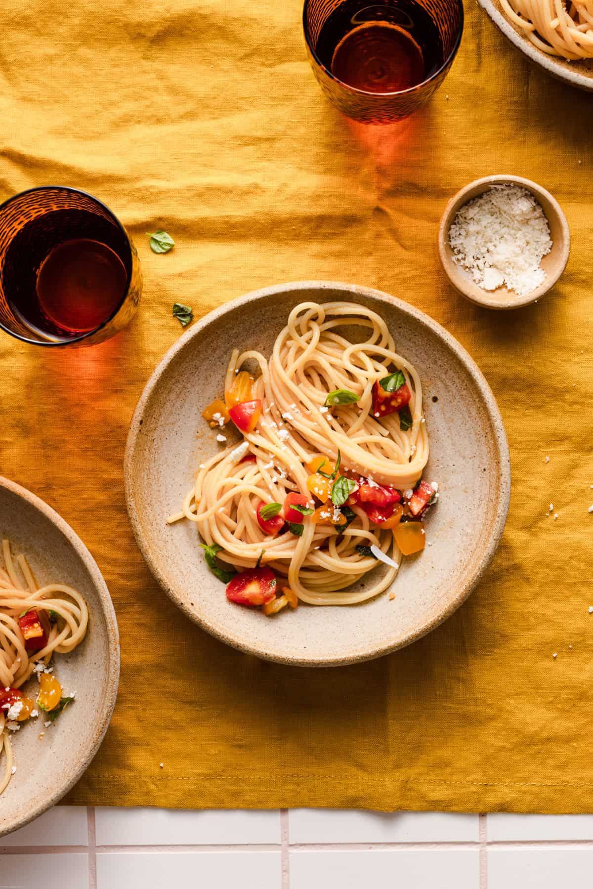 Showing the fourth step: spaghetti pasta with salsa alla crudaiola into bowls on a orange table cloth with dark orange glasses placed around and a pinch bowl covered with grated cheese.