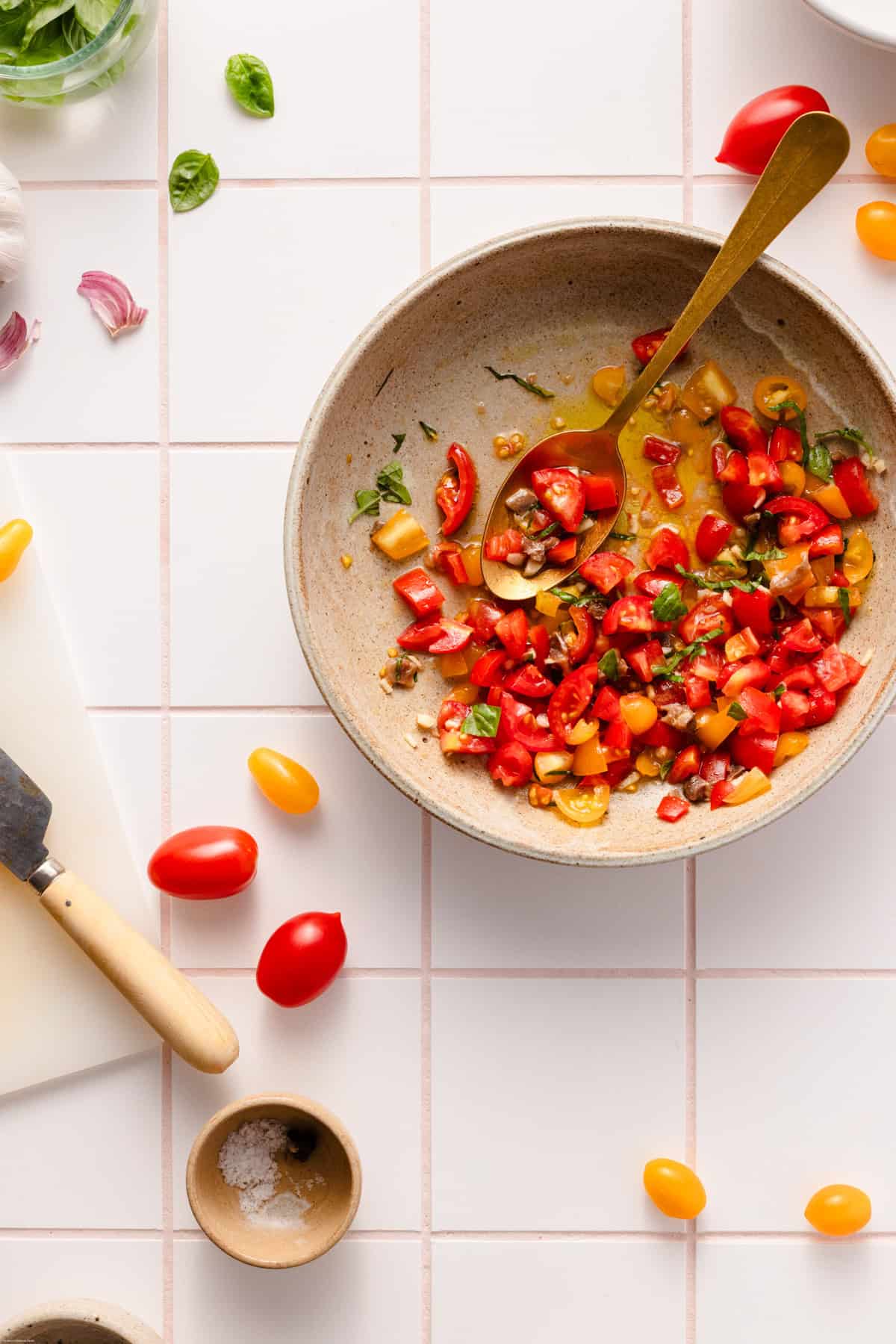 Showing how to make the second step by adding the olive oil into the big bowl with the cut tomatoes and basil. All placed on a tiles backdrops with tomatoes around.