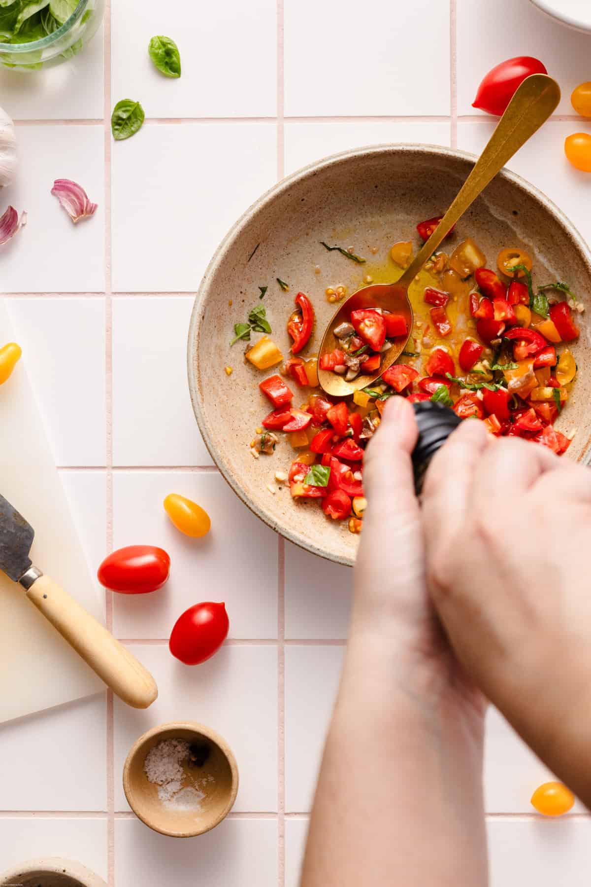 Showing how to make the third step by cracking the black pepper into the big bowl with the cut tomatoes and basil. All placed on a tiles backdrops with tomatoes around and a chopping board with knife on it.
