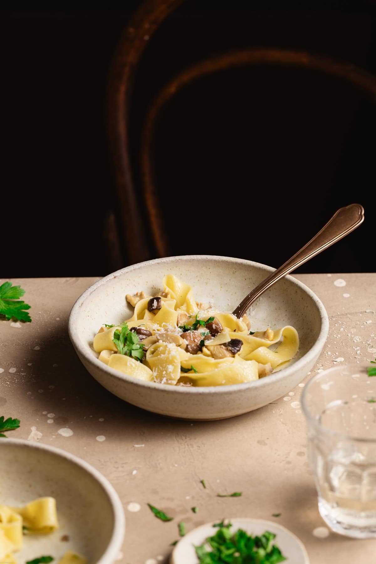 A side shot of pasta pappardelle with the mushroom sauce on a brown table next to a wine glass with parsley leafs around.