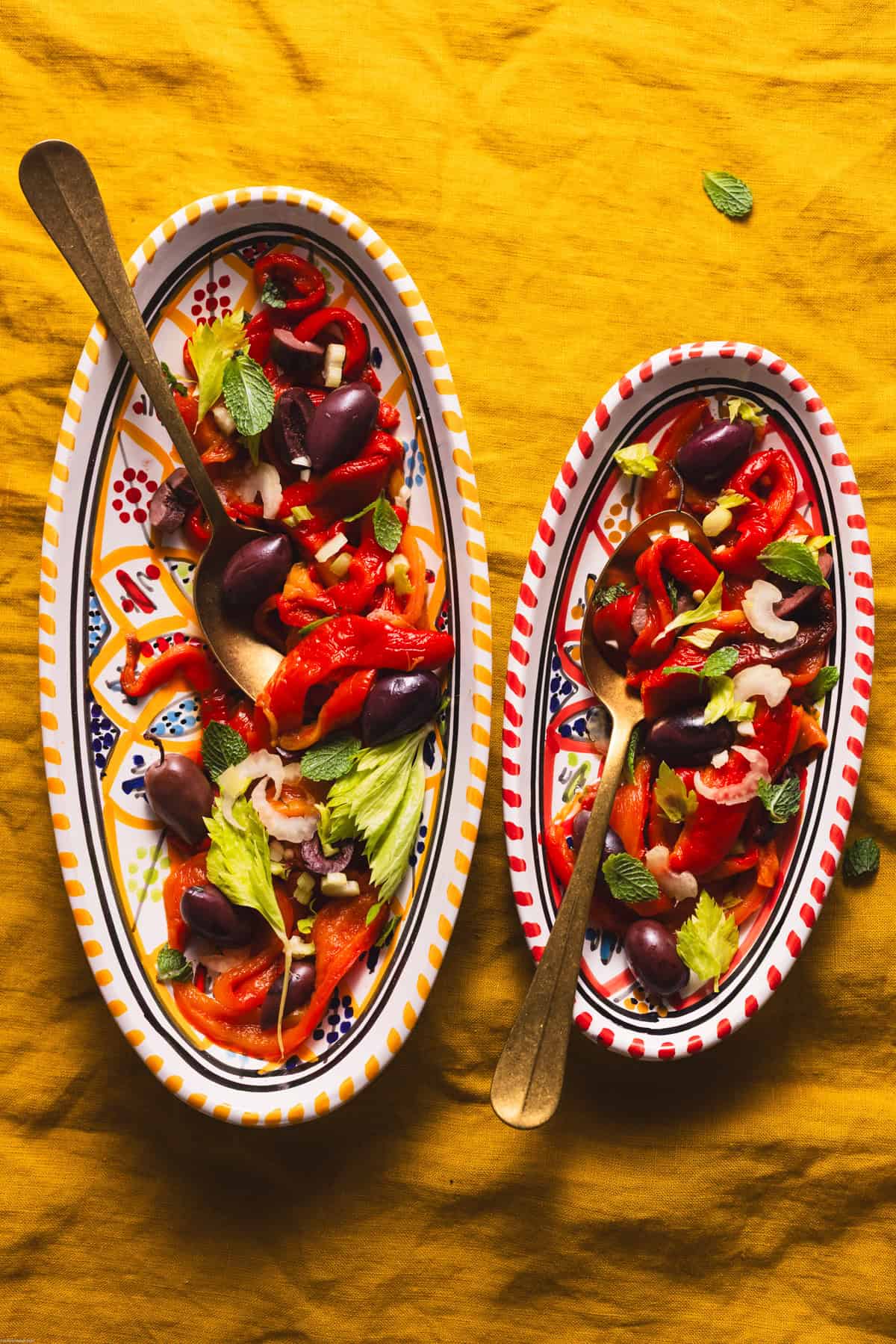 Roasted red peppers on two serving plates on a mustard linen with bronze serving spoon on it.