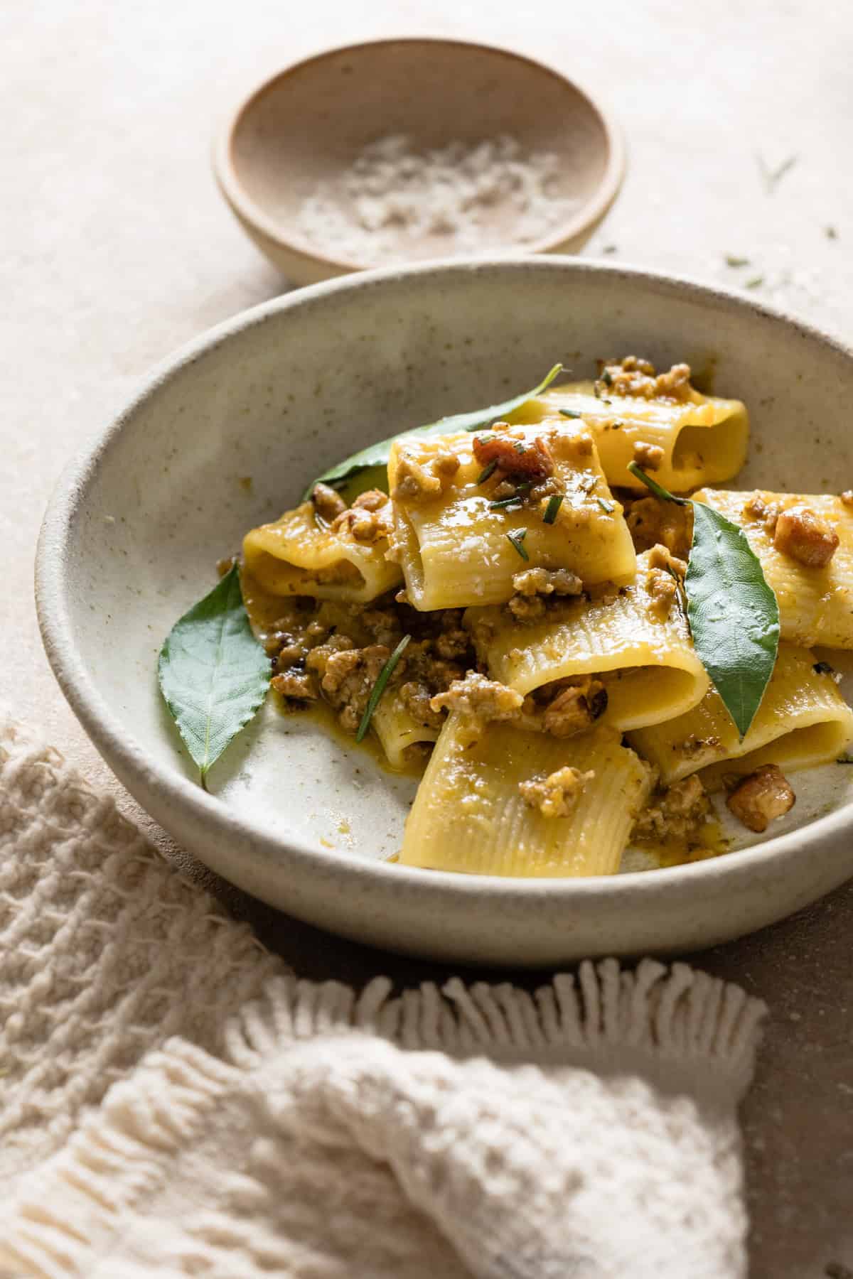 A close up side shot of white pate with ragù bianco on a white backdrop, next to a white cloth, fork, orange glass and a pinch bowl.
