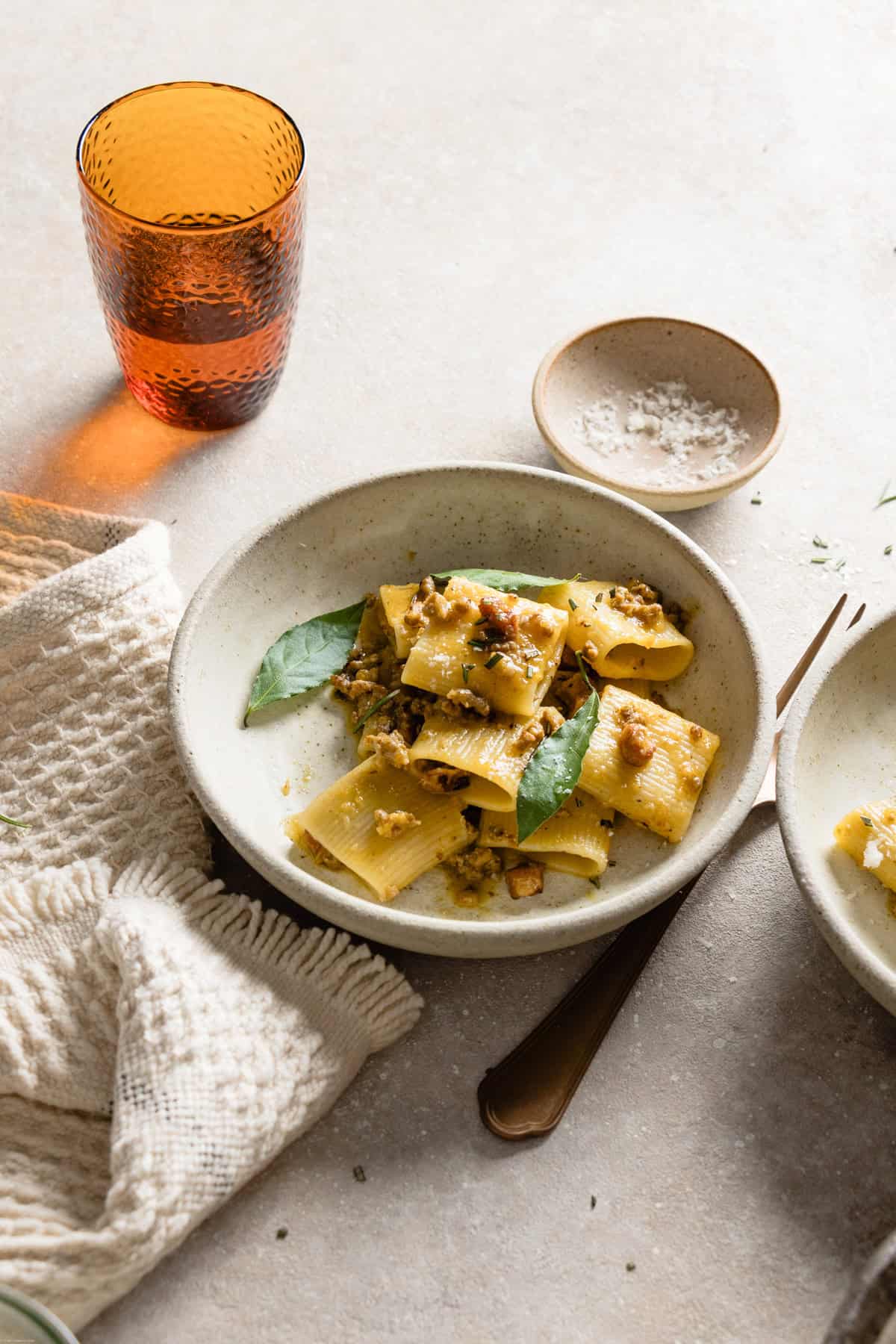 A shot of white plate with ragù bianco on a white backdrop, next to a white cloth, fork, orange glass and a pinch bowl.