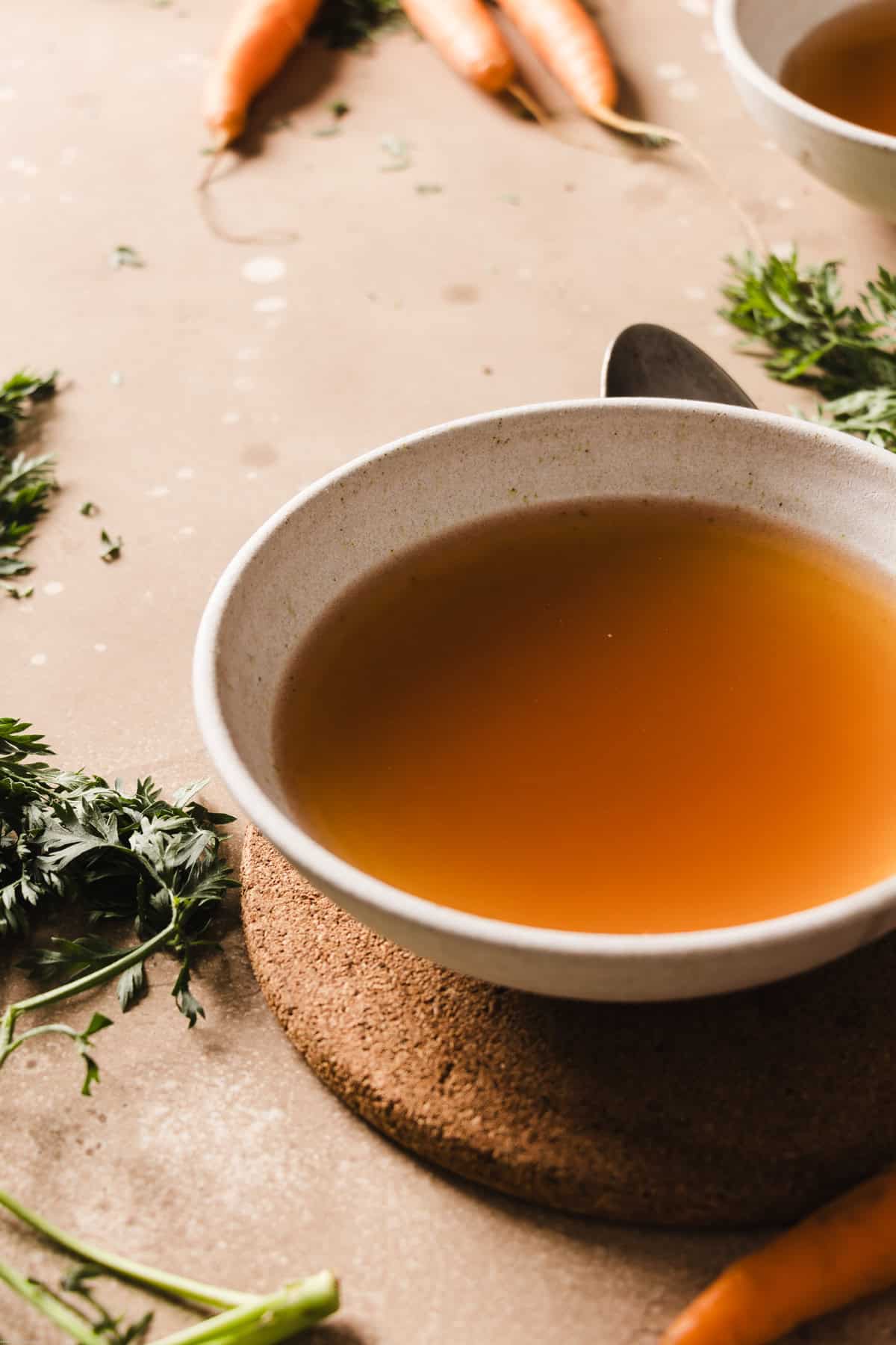 A side photo showing a bowl filled with some brodo vegetale on a brown backdrops. Around the bowl you'll find the carrots and a spoon