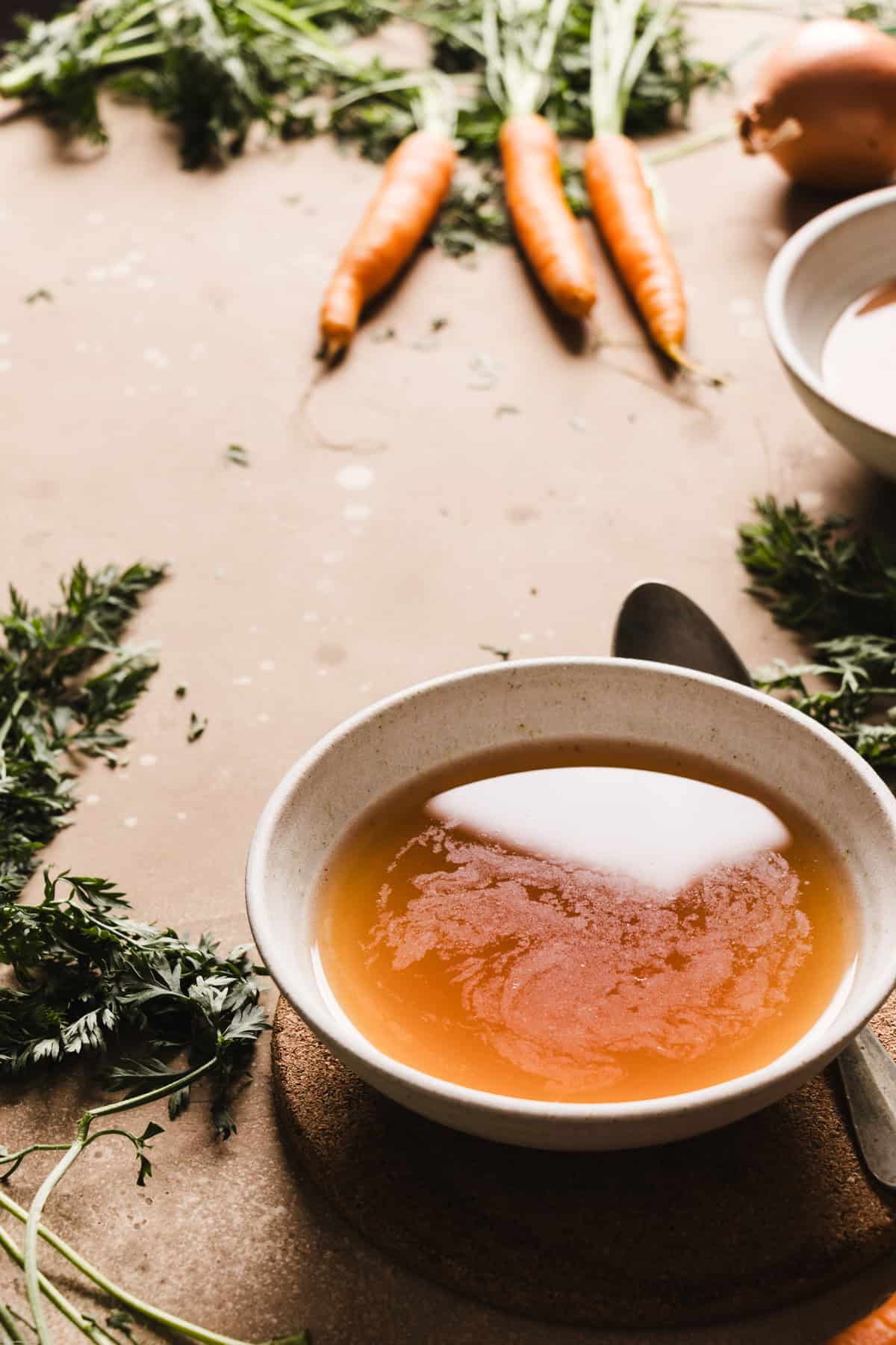 A side backlight photo showing a bowl filled with some brodo vegetale on a brown backdrops. Around the bowl you'll find the carrots and a spoon