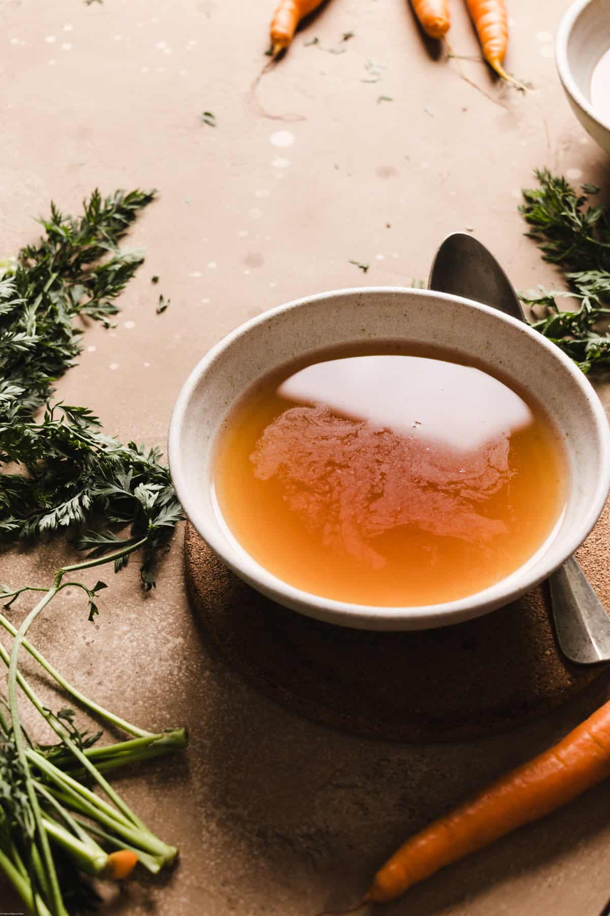 A side photo showing a bowl filled with some brodo vegetale on a brown backdrops. Around the bowl you'll find the carrots and a spoon