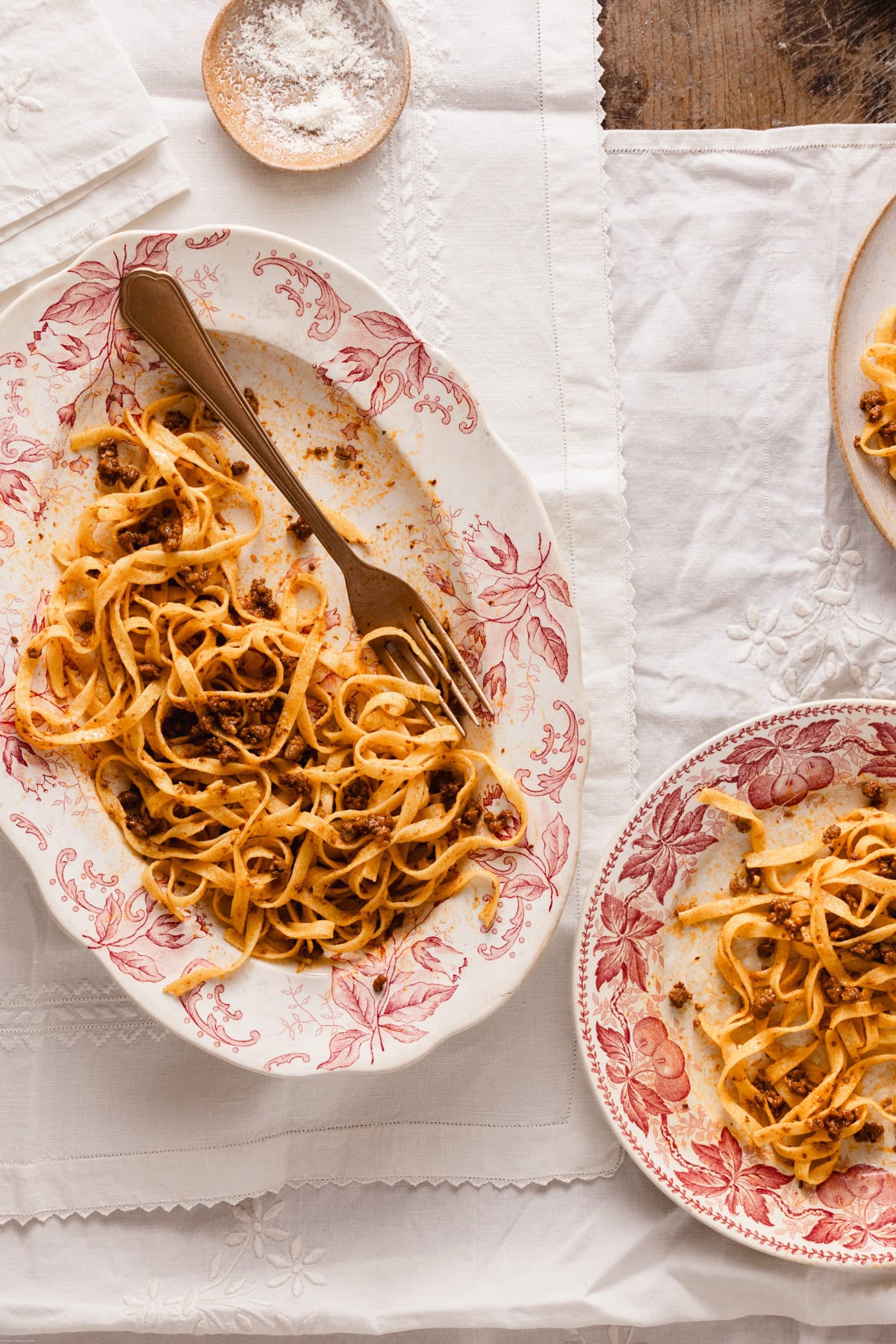 Tagliatelle on a serving plate near a plate full of pasta with ragu on a white tablecloth.