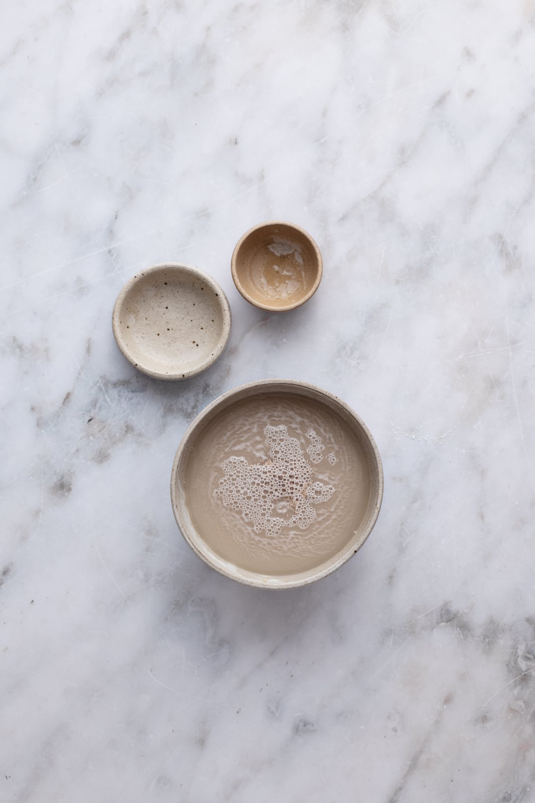 Activated yeast mixture in a ceramic bowl, ready to be added in the flour. Surrounding the bowl are small empty pinch bowls, set on a marble surface.