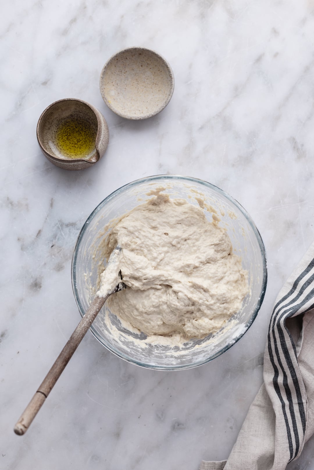 Mixed dough on a large bowl surrounded by two small bowls with olive oil and salt, set on a marble surface.